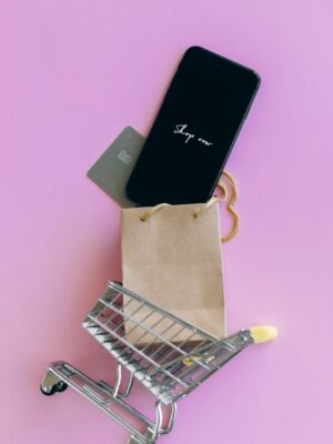 A smartphone, credit card, and bag in a miniature shopping cart on a pink background.
